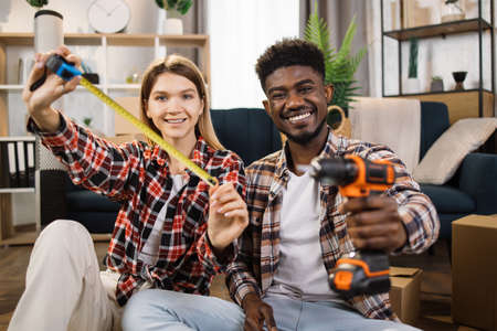 Married couple holding measure tape and electric drill while sitting in living room and smiling at camera. Handsome man and charming woman preparing to change interior at new apartment.の写真素材