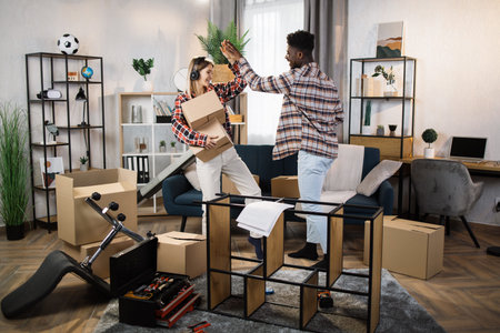 Cheerful woman in headphones giving high five to her boyfriend while holding carton boxes at bright living room. Happy multicultural couple celebrating moving day.の写真素材