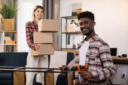 Smiling african american man assembling with drill modern furniture while caucasian woman standing behind with boxes in hands. Concept of family, moving, first home.の写真素材