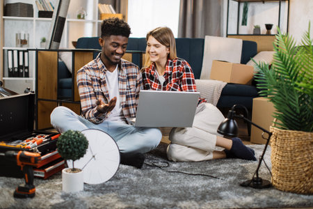 Happy young man and woman chatting and smiling while sitting on floor with wireless laptop....