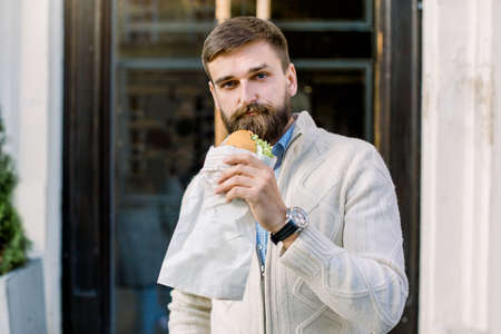 Young handsome Caucasian bearded man enjoying lunch break while holding fresh croissant standing outdoor on the background of the cafe door.の写真素材