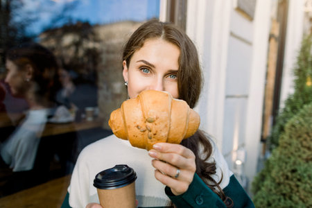 Young pretty stylish woman hiding half of her face with fresh delicious croissant and enjoying lunch...