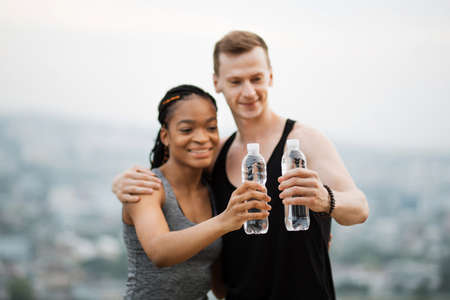 Positive multiracial couple dressed in active clothes toasting with bottle of water after outdoors training. Concept of people, sport and recreation.の写真素材