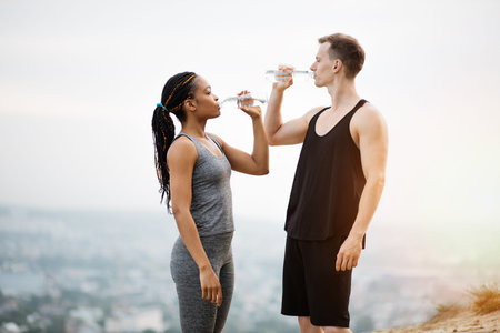 Side view of fitness young people in sport outfit drinking fresh cold water after outdoors activity. Multiracial couple leading a healthy and active lifestyles.の写真素材