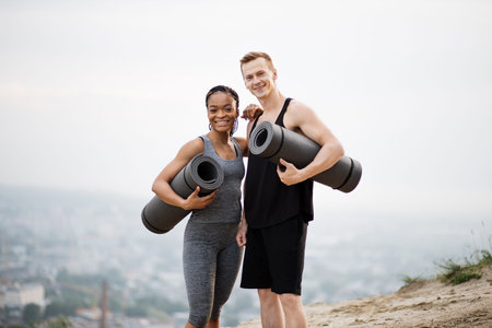 Beautiful positive multicultural couple on sport clothes holding yoga mat in hands while standing...