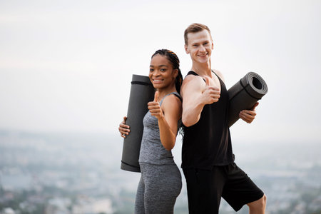 Smiling caucasian man and african woman dressed in sportswear showing thumbs up and holding yoga...