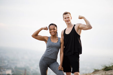 Smiling african woman and caucasian man in sport clothes showing their strong biceps while standing...