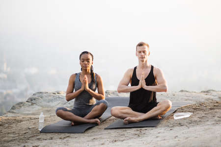 Peaceful multi ethnic couple in sport clothes sitting in lotus position yoga mat and with namaste hands. Young man and woman keeping eyes closed while meditating on fresh air.の写真素材