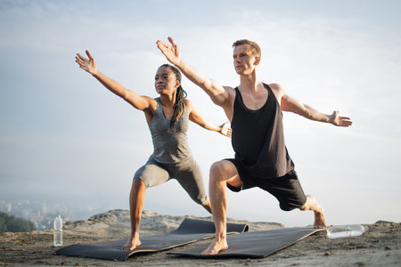 Caucasian man and african american woman dressed in activewear standing in with bare feet on yoga mat and exercising together with arms outstretched. View of big city among clouds on background.の写真素材