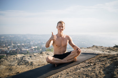 Smiling joyful handsome caucasian man with bare torso sitting in lotus position on yoga mat and...
