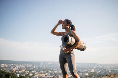 Fitness black woman in activewear holding yoga mat and water bottle outdoors. Slim young female standing on hill and looking aside. Cityscape in background.の写真素材