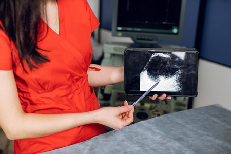 Close up cropped horizontal image of female doctor in red uniform, holding digital tablet pc and pointing with the pen on the monitor with ultrasound scan of the abdomen and kidney.の写真素材