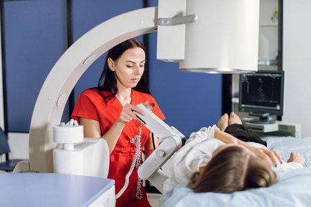 Young attractive focused woman doctor in red uniform, providing lithotripsy procedure for her lying female patient with modern ultrasonic lithotriptor to break up stones.の写真素材