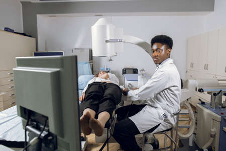 Extracorporeal shock wave therapy. Kidney stone treatment. Front view of African male doctor looking at the digital screen of the lithotriptor to determine stones position of woman patient.の写真素材