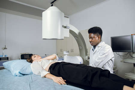 Young high-skilled focused black man doctor in white coat, providing lithotripsy procedure for his lying female patient with modern ultrasonic lithotriptor to break up stones.の写真素材