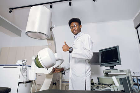 Portrait of handsome young professional African man doctor standing near modern ultrasound and lithotripter machines and showing thumb up. Safety non-invasive stone treatment, shock wave lithotripsy.の写真素材