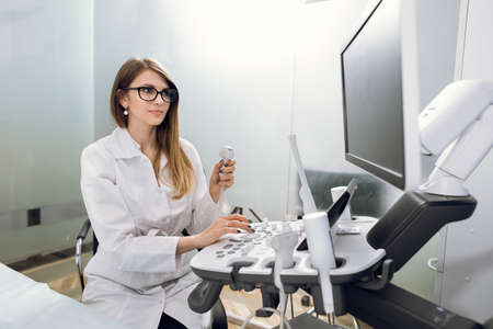 Young female Caucasian doctor working on modern ultrasound equipment. Operator of ultrasound scanning machine sitting and looking at the monitor, waiting for patientの写真素材