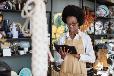 Pretty afro american woman in eyegasses using digital tablet for making inventory at decor shop. Attractive saleswoman in apron standing with modern gadget in hands.の写真素材