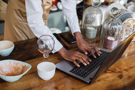 Close up of african saleswoman in beige apron using laptop at store during receipt of goods. Beautiful various decor at shop. Sale concept.の写真素材
