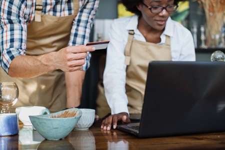 Close up of african woman and caucasian man using credit card and modern laptop while working at decor store. Concept of people, sale and coworking.の写真素材