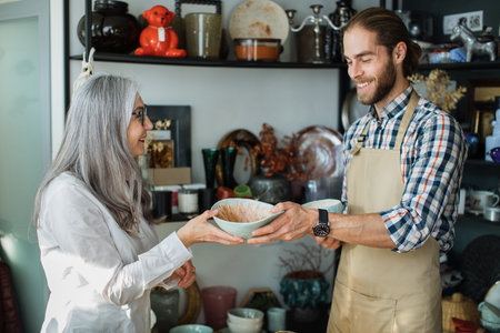 Caucasian bearded seller in beige apron consulting mature woman at decor store. Shelves with various exclusive goods. Concept of shopping and sale.の写真素材