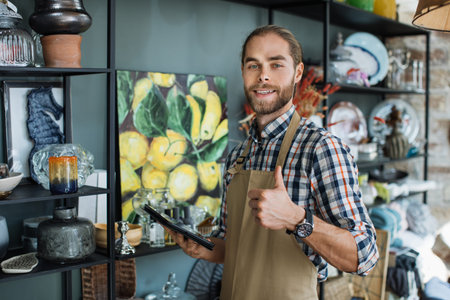 Smiling young man showing thumb up and holding digital tablet while standing at modern store with various decor. Concept of service, goods and shopping.の写真素材