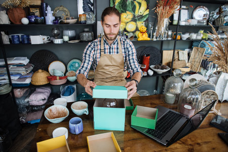 Handsome caucasian seller receiving unique goods at decor shop. Bearded man in beige apron sitting...