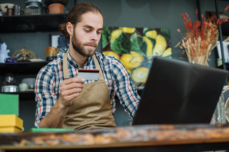 Bearded salesman using wireless laptop at decor store for registration discount card of regular customer. Modern service and loyalty at shop.の写真素材