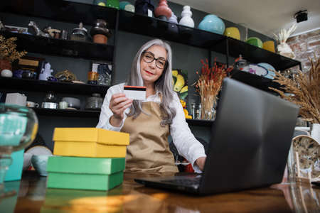 Smiling 50-aged gray-haired woman decor shop worker, sitting at counter, typing on modern laptop and holding credit card in hand. Online payment, shopping concept.の写真素材
