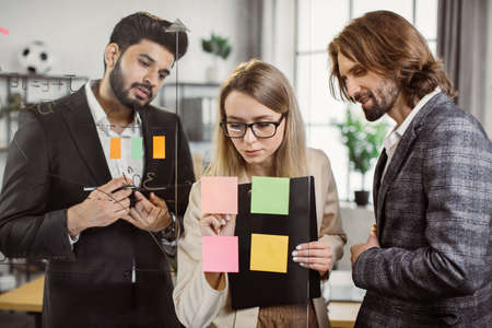 Group of multicultural professional brokers sticking notes with project information on glass wall during office meeting. Partners with tablet and clipboard cooperating together at modern workplace.の写真素材