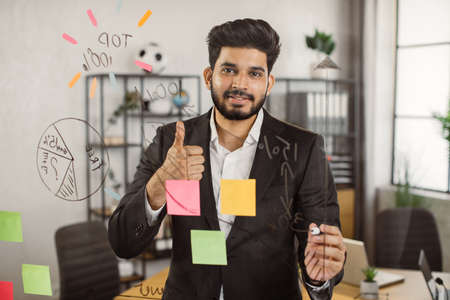 Portrait of executive male manager showing thumb up sign while working on new project at office. Smiling indian man standing in front of glass board and sharing with motivation mood.の写真素材