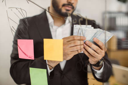 Loose up of concentrated male chief in formal clothes counting money while standing in front of glass board at own company. Salary and business earnings concept.の写真素材