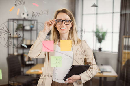 Happy caucasian project manager with blond hair posing with toothy smile in front of glass wall at boardroom. Attractive woman in eyeglasses using digital tablet while working at creative office.の写真素材