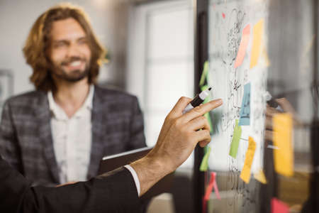 Close up of businessman holding felt-tip pen and pointing on interactive transparent board with written diagrams while his colleague staining near. Concept of business, people and startup.の写真素材