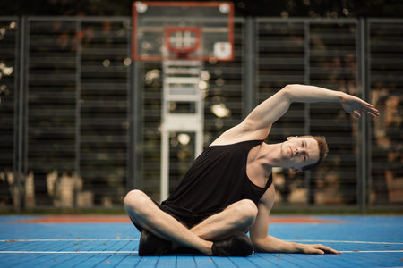 Happy fitness sporty man in black sportswear stretching his arms and warming up while sitting on the...