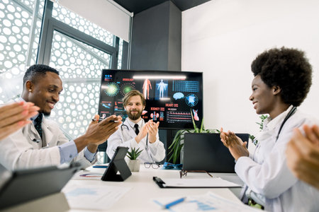 Multiethnic team of five doctors at the table, clapping their hands, applauding and enjoy successful...