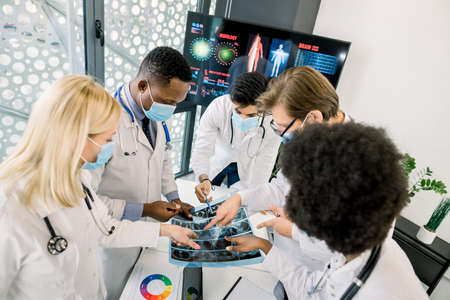 Top angle view of multiethnical team of doctors in medical masks, examining CT scan together, discussing diagnosis and ways of successful treatment. Medical workers looking at scan at the hospital.の写真素材