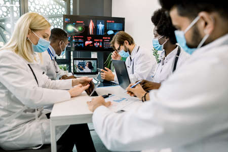 Group of multiethnical healthcare workers in masks, with digital tablets and laptop, meeting in hospital boardroom. Medical staff during morning online briefing with their chief doctor, African lady.の写真素材