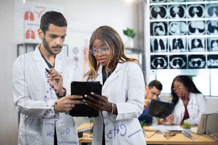 Two multiethnic male and female doctors standing behind glass wall with MRI scan and working on tablet pc. Their colleagues talking each other at the table on backgroundの写真素材