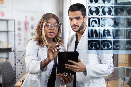 Two multiracial doctors using tablet pc while examining tomography of patient during consilium....
