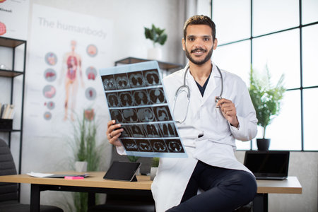 Portrait of smiling likable Arabian Malay male doctor radiologist, leaning on the table in modern clinic office, and holding an x-ray tomography scan.の写真素材