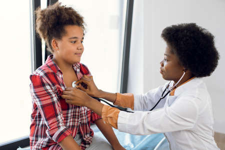 African female pediatrician with stethoscope auscultating cute teen mixed race girl patient. Black physician, checks heartbeat and lungs breathing of kid, making checkup before vaccinationの写真素材
