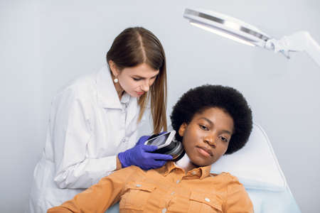 Young African American woman client lying on the couch in modern dermatology clinic, having face skin diagnostic. Professional young female dermatologist studying skin type using dermatoscopeの写真素材