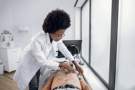 Medicine, health and heart check-up concept. Young afro-american woman doctor with electrocardiogram equipment making cardiogram test to male patient in hospital, fixing electrodes on patient chestの写真素材