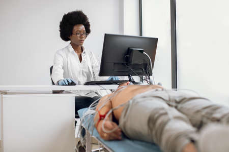 Young focused African American woman doctor, sitting at the table and working on computer, while performing cardiology ECG test for male patient in hospital emergency roomの写真素材