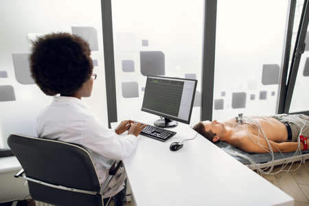 Back view of female African doctor cardiologist, looking on the computer screen on electrocardiogram results of young man. Man patient lies on the couch. Diagnosis and treatment of heart attackの写真素材