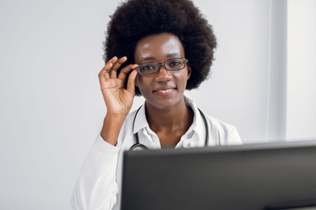 African-American doctor woman, sitting at the table in modern clinic, working on laptop pc, giving...