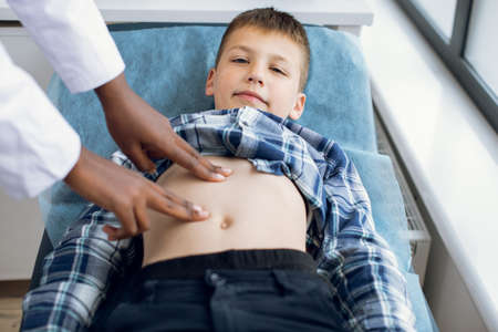 Cute smiling 10-aged kid boy, lying on the couch at modern clinic, visiting his doctor. Female afro pediatrician examining abdomen of child patient using manual palpationの写真素材