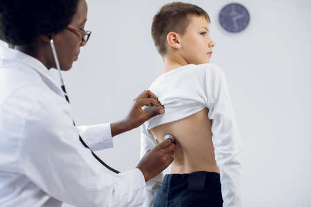 Respiratory disease, asthma and pneumonia diagnostics and treatment. Close up cropped shot of female african doctor, examining teen 10-aged schoolboy patient with stethoscopeの写真素材
