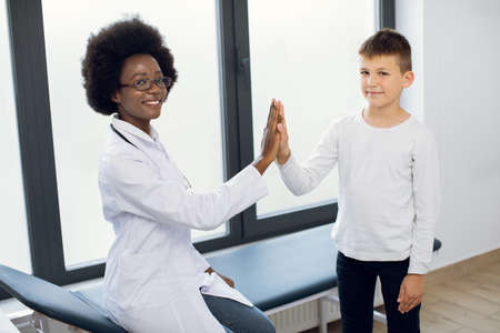 Young pretty likable medical worker, Afro-American lady doctor gp pediatrician, giving her cute child boy patient a high five after successful annual medical check up in modern clinicの写真素材